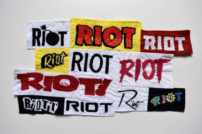 Eleven fabric rectangles pieced together in an uneven shape. Each one features the word ‘riot’ hand embroidered in different fonts, colours and sizes.